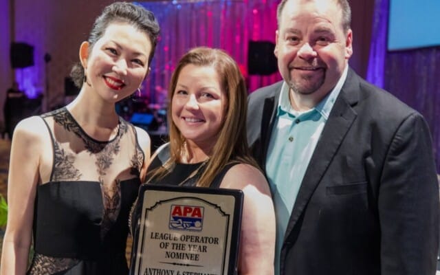 Three people smiling, one holding a plaque reading "APA League Operator of the Year Nominee". Their dedication to the American Poolplayers Association shines through in their joyful expressions.