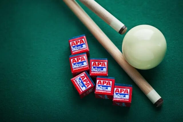 Pool sticks, a cue ball, and several red chalk cubes featuring the "APA" branding are scattered on a green felt surface, capturing the essence of the American Poolplayers Association.
