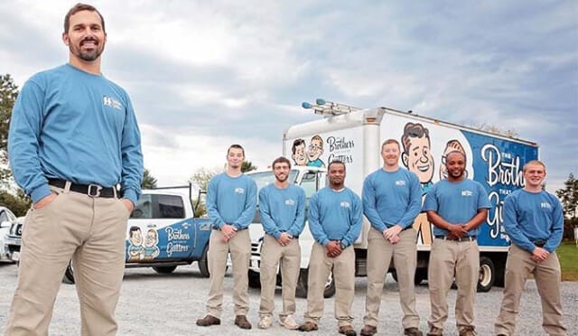 A team of service professionals from The Brothers That Just Do Gutters franchise posing in front of their company vehicles with the company's logo and branding visible.