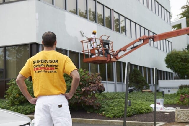 A man in a yellow "supervisor" shirt from CertaPro Painters observes a cherry picker outside a modern office building.