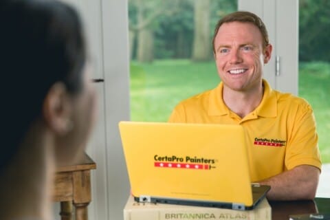 A smiling man in a yellow CertaPro Painters shirt using a laptop with the CertaPro Painters logo, sitting across from another person at a table.