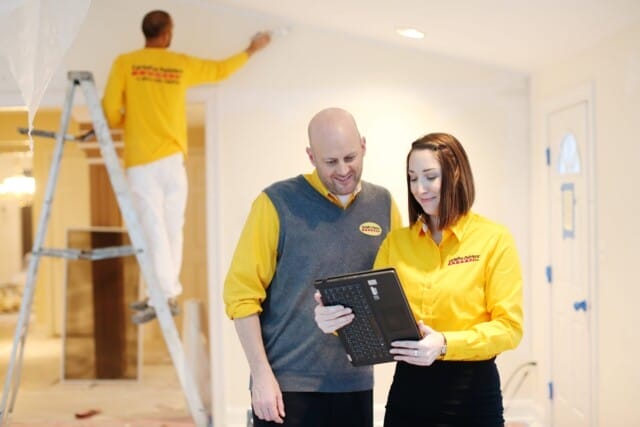 Two professional painters in yellow CertaPro shirts review a laptop while another painter works on a ladder in the background.
