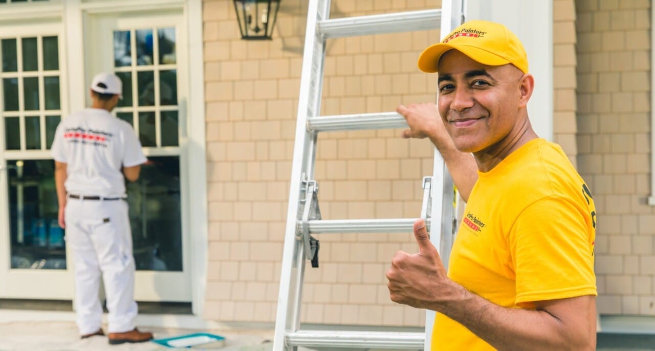 A smiling man in a yellow shirt and hat giving a thumbs-up beside a ladder, with another professional painter working in the background by a house.