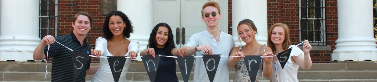 Group of six smiling young adults from Class-101 holding a banner that spells "Seniors" in front of a building with columns.