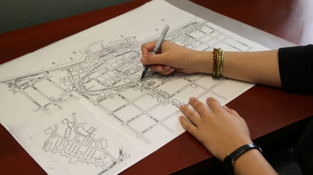 Person's hands using a pen to work on a detailed Discovery Map laid on a table, with additional sketches nearby.