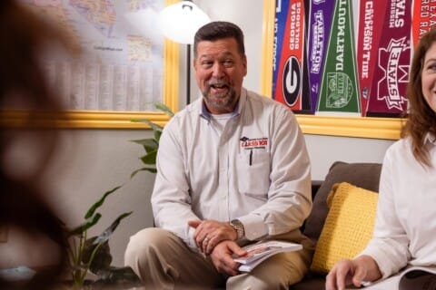 A leader from Class 101 seated on a couch in a room with maps on the wall training franchisees.