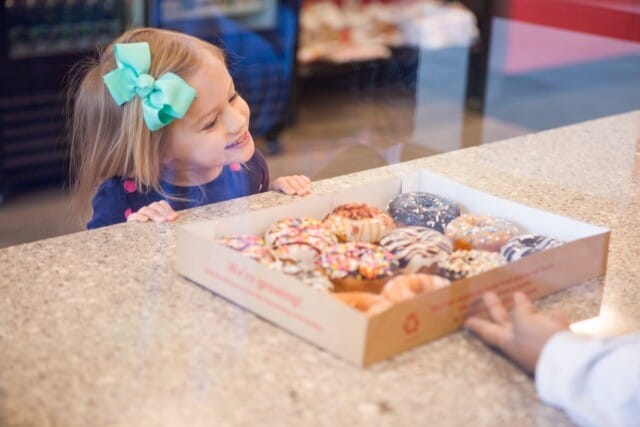 A young girl with a large bow in her hair excitedly looks at a box of various colorful Duck Donuts on a counter.