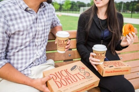 Two people sitting on a park bench, enjoying coffee and Duck Donuts from branded containers, with a focus on the food and their smiles.