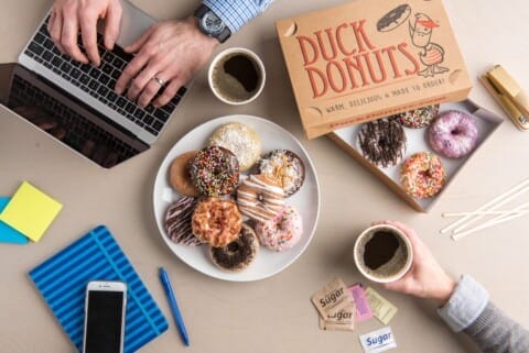 A top-down view of a work desk with two people, assorted specialty donuts from Duck Donuts on a plate, a laptop, coffee cups, a smartphone, and office supplies.