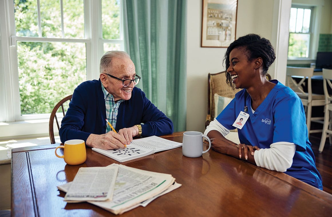 Elderly man doing a crossword puzzle at a table with a smiling healthcare worker from BrightStar Care franchise.