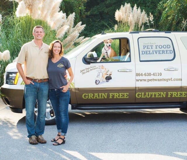 Two Pet Wants franchisees stand smiling next to a branded van. A dog looks out from the driver's seat.