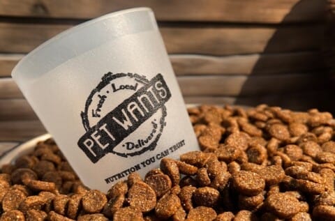 Plastic cup labeled "Pet Wants" on top of a pile of brown pet food kibble with a wooden background.