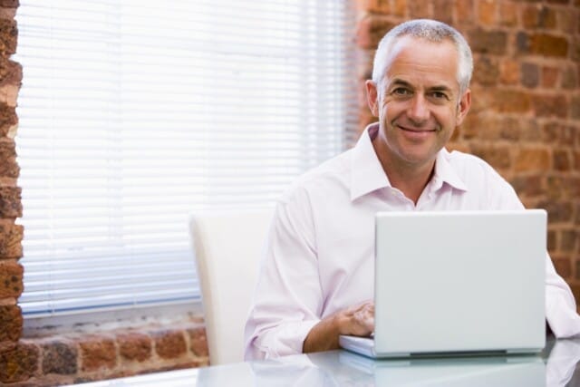Smiling franchisee in a white shirt working on a laptop in an office with brick walls.