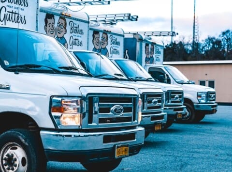 A lineup of white service trucks parked in a row, each adorned with "The Brothers That Just Do Gutters" branding.