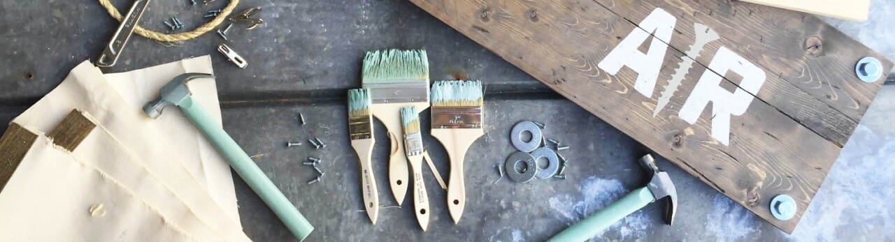 Craft materials and tools including paintbrushes, hammers, screws, washers, rope, and a wooden board with "AR" stenciled on it are arranged on a surface.