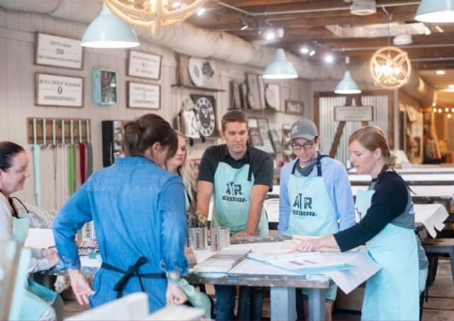 Group of franchisees undergoing training at AR Workshop wearing aprons, gathered around a table with art and craft materials.