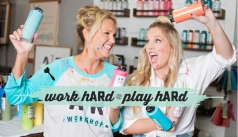 Two AR Workshop franchisees smiling and playfully holding paint bottles in a colorful workshop setting.
