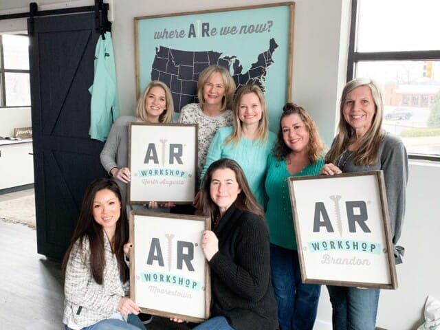 Group of six smiling women holding signs for different AR Workshop locations, standing indoors with a map in the background.