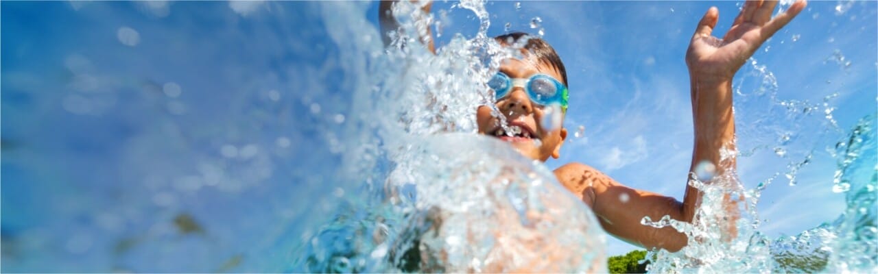 Young boy wearing goggles swimming in a pool and splashing towards the camera.