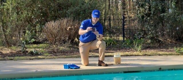 A man in a blue uniform representing America's Swimming Pool Company, kneels by a poolside, holding a water testing kit.