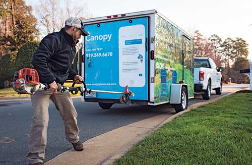 A worker carrying a leaf blower walks past a Canopy Lawn Care-branded service vehicle parked on a suburban street.