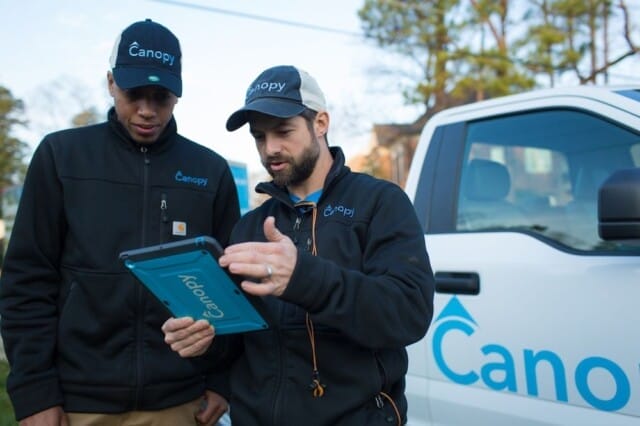 Two technicians in branded Canopy Lawn Care workwear using a tablet beside their franchise company van.