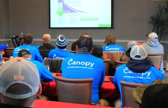 Group of people wearing Canopy Lawn Care branded apparel attending a franchise training presentation.