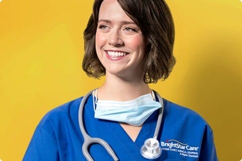 A smiling healthcare professional in blue scrubs with a stethoscope and a partially lowered face mask, completing their training at a franchise location.