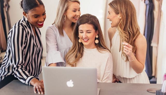 Four women smiling and looking at a laptop screen together in a bright room for Bella Bridesmaids franchise training.