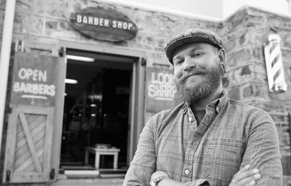 Bearded franchise owner standing in front of a barber shop with arms crossed.