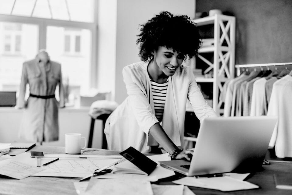 Fashion designer working on a laptop in a studio with sketches and patterns on the desk.