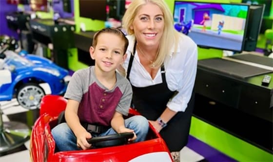 A child sits in a toy car at a brightly colored play area, as they prepare for their fun experience at Sharkey's Cuts for Kids..