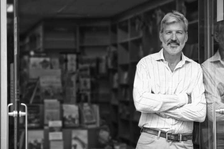 Middle-aged franchise owner smiling with crossed arms in front of his bookstore, offering his business for sale.