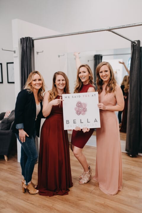 Four women smiling and holding a sign that reads "we said yes at Bella Bridesmaids" in a bridal shop.