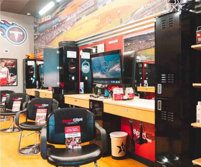 The interior of Sport Clips boasts a vibrant sports-themed decor with TVs to catch the game, sleek hair cutting stations, black chairs accented in red, and lockers filled with sports memorabilia.