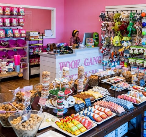 Woof Gang Bakery interior featuring pink walls, snacks on display tables, and toys on shelves.